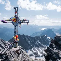 Croix ornée de rubans au sommet rocheux du massif du Carlit, dominant des montagnes bleutées sous un ciel nuageux.