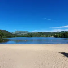 Plage de sable fin au bord d'un lac paisible, avec des montagnes verdoyantes en arrière-plan, sous un ciel bleu éclatant. Le Puy de Sancy veille sur ce paysage idyllique.