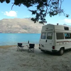 Camping-car blanc au bord d'un lac turquoise, table et chaises dressées. Un décor parfait pour un budget nouvelle zélande.