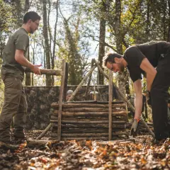 Apprendre comment faire du feu dans la nature : des hommes construisent une structure en bois pour un feu de camp, avec un pot suspendu.