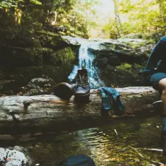 Un randonneur se repose près d'une cascade, ses bottes et chaussettes posées sur une bûche. Une pause pour réfléchir à comment éviter de transpirer des pieds.