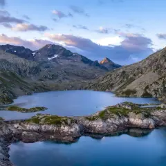 Vue aérienne du lac des 7 Laux, entouré de montagnes rocheuses et verdoyantes, sous un ciel aux teintes rosées.