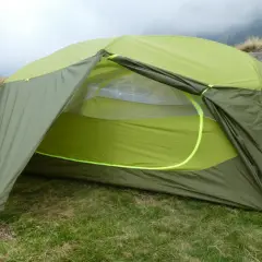 Tente verte et jaune, équipement de camping Nemo, installée sur une pelouse avec des montagnes brumeuses en arrière-plan.