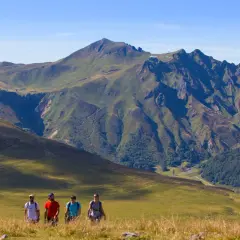 Quatre randonneurs marchent dans un paysage de montagne verdoyant, avec le plus haut sommet du Massif Central en toile de fond.