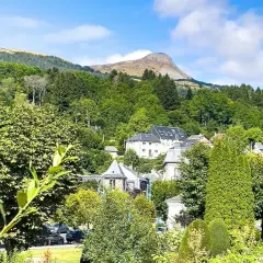 Vue d'un village niché dans une vallée verdoyante, avec des maisons aux toits gris et un grand bâtiment blanc. Au loin, une montagne imposante se dresse sous un ciel bleu. Un banne ordanche, peut-être une tente ou un auvent, est visible dans le paysage.