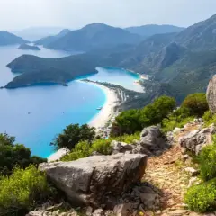 Sentier rocailleux du Lycian Way surplombant une baie turquoise et une plage de sable blanc bordée de montagnes verdoyantes.
