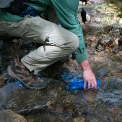 Randonneur remplissant une gourde dans un ruisseau, prêt à utiliser une pastille de chlore pour eau potable.