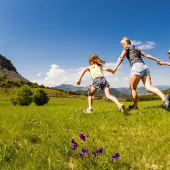 Une mère et ses deux filles courent main dans la main dans un champ fleuri, avec le Mont Gerbier de Jonc en toile de fond.