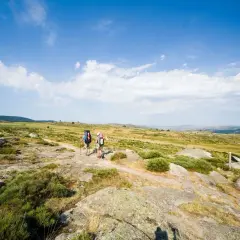 Deux randonneurs avancent sur un sentier rocailleux, sous un ciel bleu parsemé de nuages. Ils parcourent les gr 70 étapes, profitant du paysage.
