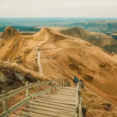 Une personne entame une rando au Puy de Sancy, descendant un escalier en bois sur un flanc de montagne aux herbes dorées.