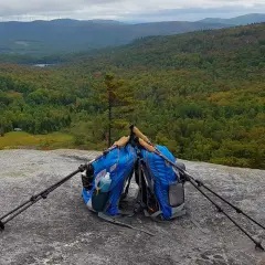 Deux sacs de rando bleus sont posés sur un rocher, avec des bâtons de marche plantés à côté. Le paysage vallonné et boisé s'étend à l'horizon.