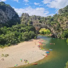 Les gorges du Chassezac, un pont naturel rocheux surplombe une rivière verdoyante où des kayakistes profitent du soleil sur une plage de galets.