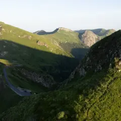 Un randonneur solitaire au sommet d'une colline verdoyante, dominant un paysage montagneux spectaculaire. Une route sinueuse serpente dans la vallée. Randonnée Cantal.
