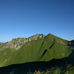 Vue panoramique sur des montagnes verdoyantes sous un ciel bleu clair. Un sentier sinueux suggère une randonnée de **gr30 difficulté**.