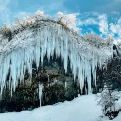 Impressionnante cascade gelée en Auvergne en hiver, avec des stalactites de glace bleutée tombant d'une falaise enneigée sous un ciel bleu et nuageux.