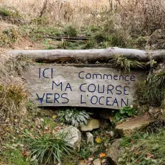 Panneau en bois aux **sources de la Loire** : "Ici commence ma course vers l'océan". Entouré d'herbes sèches et de rochers.