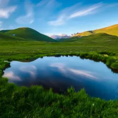 Un étang reflète le ciel et les collines verdoyantes, évoquant la petite Mongolie auvergnate. Un paysage paisible avec des nuages filant.
