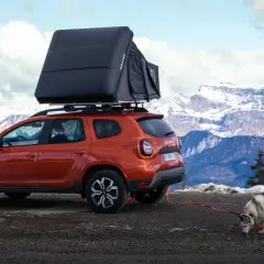 Un Dacia Duster orange avec tente de toit, prêt pour un bivouac Vanoise. Un husky renifle le sol, la neige et les montagnes en arrière-plan.