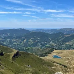 Vue panoramique des montagnes verdoyantes du Cantal, avec un lac bleu scintillant au loin. Le **Plomb du Cantal** domine l'horizon sous un ciel bleu parsemé de nuages.