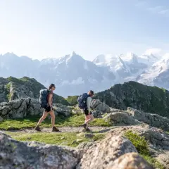 Deux randonneuses avec sacs à dos traversent un sentier rocheux, avec des montagnes enneigées en arrière-plan. Une randonnée accessible en train pour admirer ce paysage.