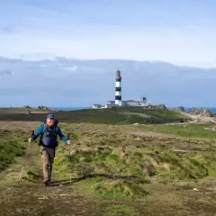 Un randonneur souriant marche sur un sentier côtier à Ouessant, avec le phare en arrière-plan.