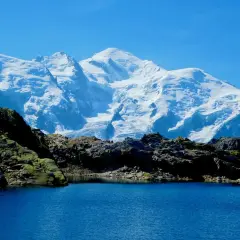 Lac d'altitude aux eaux bleues, bordé de roches sombres, avec en arrière-plan le massif enneigé du Mont-Blanc, paysage typique du trek des Aiguilles Rouges.