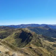 Vue panoramique sur les montagnes du puy Cantal, avec des crêtes herbeuses et des forêts verdoyantes sous un ciel bleu éclatant.