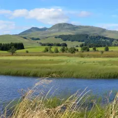 Vue paisible du lac Chauvet, entouré de prairies verdoyantes et de collines ondulantes sous un ciel bleu parsemé de nuages.