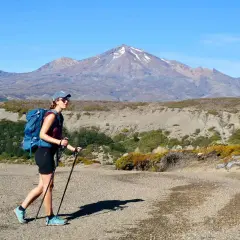 Randonneuse avec sac à dos et bâtons, portant une chaussure sans chaussette, marche sur un sentier rocailleux devant une montagne enneigée.