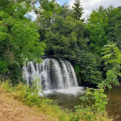 Belle cascade auvergne, entourée de verdure luxuriante. L'eau tombe en rideau sur des rochers, rejoignant une rivière paisible. Un bâtiment ancien se dresse à droite.