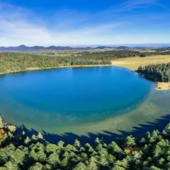 Vue aérienne du lac de Servières, entouré de forêts verdoyantes et de montagnes bleutées sous un ciel clair.