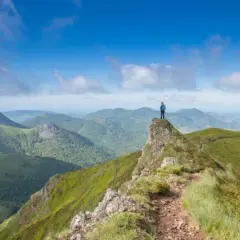 Un randonneur au sommet d'une crête verdoyante, contemplant un panorama de montagnes. Quel est le plus beau coin du Cantal ? Ce paysage spectaculaire en est un candidat.