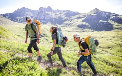 Trois randonneurs équipés de gros sacs à dos, dont un avec une corde, gravissent une pente verdoyante. La laine vierge, comme celle de leurs vêtements, est idéale pour l'aventure.