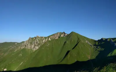 Vue panoramique sur des montagnes verdoyantes sous un ciel bleu clair. Un sentier sinueux suggère une randonnée de **gr30 difficulté**.