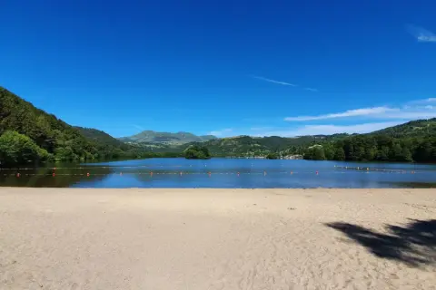 Plage de sable fin au bord d'un lac paisible, avec des montagnes verdoyantes en arrière-plan, sous un ciel bleu éclatant. Le Puy de Sancy veille sur ce paysage idyllique.