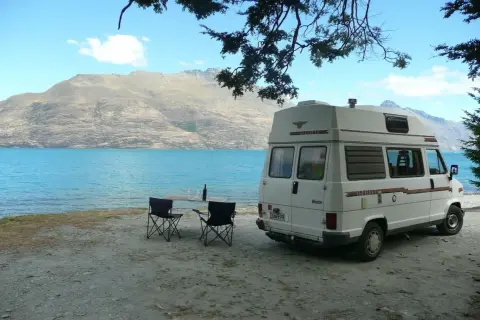 Camping-car blanc au bord d'un lac turquoise, table et chaises dressées. Un décor parfait pour un budget nouvelle zélande.