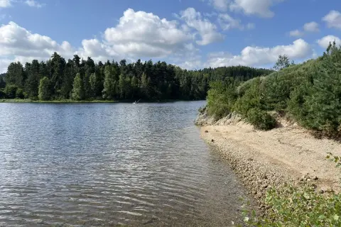 Magnifique paysage pour un tour du lac de Naussac à vélo. Eau calme, plage de galets et forêt dense sous un ciel bleu et nuageux.