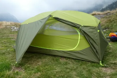 Tente verte et jaune, équipement de camping Nemo, installée sur une pelouse avec des montagnes brumeuses en arrière-plan.