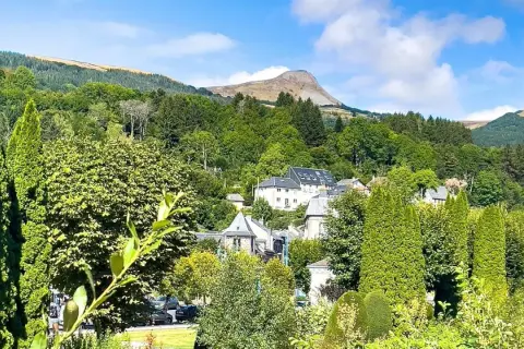 Vue d'un village niché dans une vallée verdoyante, avec des maisons aux toits gris et un grand bâtiment blanc. Au loin, une montagne imposante se dresse sous un ciel bleu. Un banne ordanche, peut-être une tente ou un auvent, est visible dans le paysage.