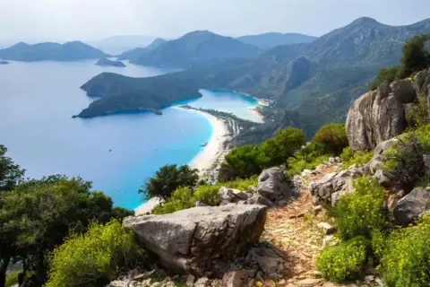Sentier rocailleux du Lycian Way surplombant une baie turquoise et une plage de sable blanc bordée de montagnes verdoyantes.