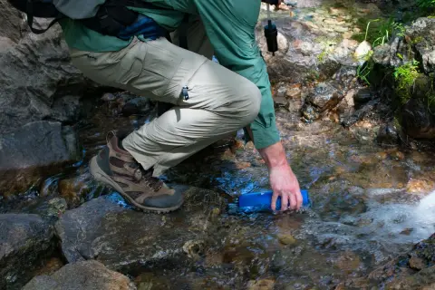 Randonneur remplissant une gourde dans un ruisseau, prêt à utiliser une pastille de chlore pour eau potable.