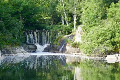 Belle cascade au lac de Naussac, entourée de verdure luxuriante. L'eau tombe sur des rochers, créant une mousse blanche.