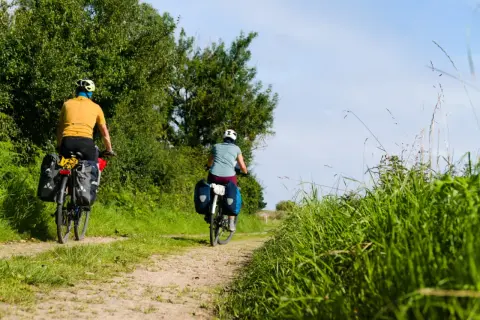 Deux cyclistes explorent un chemin de terre bordé d'arbres et d'herbes hautes. Le soleil brille, un décor idyllique pour le meilleur endroit pour faire du vélo en France.