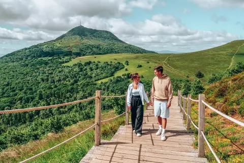 Couple se promenant sur un sentier en bois, admirant les plus beaux endroits d'Auvergne, avec le Puy de Dôme en arrière-plan.