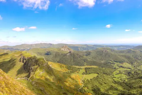 Magnifique panorama du Puy Mary, avec ses crêtes verdoyantes et ses vallées boisées sous un ciel bleu éclatant.