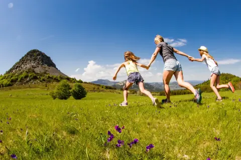 Une mère et ses deux filles courent main dans la main dans un champ fleuri, avec le Mont Gerbier de Jonc en toile de fond.
