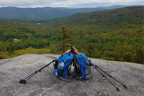 Deux sacs de rando bleus sont posés sur un rocher, avec des bâtons de marche plantés à côté. Le paysage vallonné et boisé s'étend à l'horizon.