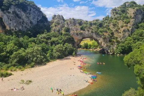 Les gorges du Chassezac, un pont naturel rocheux surplombe une rivière verdoyante où des kayakistes profitent du soleil sur une plage de galets.