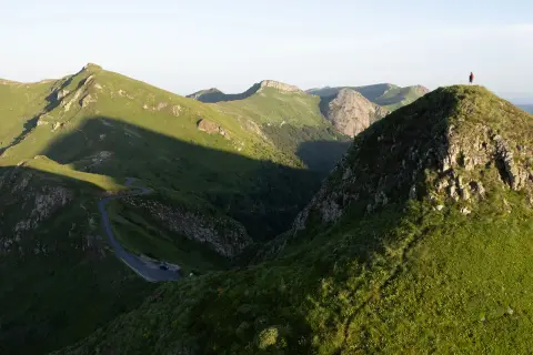 Un randonneur solitaire au sommet d'une colline verdoyante, dominant un paysage montagneux spectaculaire. Une route sinueuse serpente dans la vallée. Randonnée Cantal.