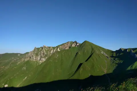 Vue panoramique sur des montagnes verdoyantes sous un ciel bleu clair. Un sentier sinueux suggère une randonnée de **gr30 difficulté**.